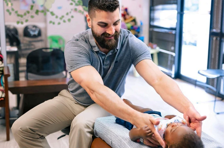 A practitioner is providing a hands-on chiropractic adjustment to a baby lying on a treatment table in a clinic setting.